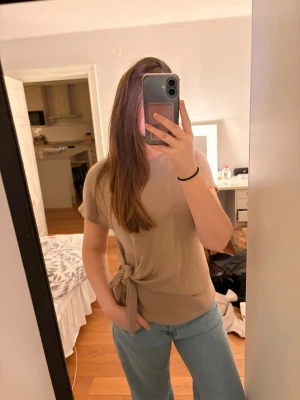 Beige topp med knyt från Zara - Säljer en beige topp från Zara med korta ärmar och rund hals. Toppen har en snygg knyt-detalj i sidan som ger en unik look. Materialet känns mjukt och lätt, perfekt till jeans eller kjol.