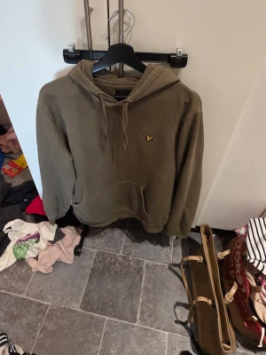 Olivgrön hoodie från Lyle & Scott - Säljer en olivgrön hoodie från Lyle & Scott med klassisk gul logga på bröstet. Hoodien har huva med dragsko och en stor magficka framtill. Tillverkad i mjuk bomullsblandning och har ribbade muddar vid ärmar och nederkant.