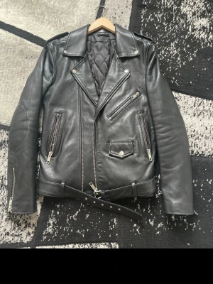 Svart skinnjacka från Acne Studios - Säljer en klassisk svart skinnjacka från blk dnm med breda slag, flera dragkedjeförsedda fickor och bälte i midjan. Jackan har quiltat foder och cool bikerstil som ger en edgy look. Perfekt för dig som vill ha en tidlös och trendig jacka.