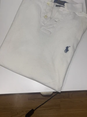 Vit pikétröja från Ralph Lauren - Klassisk vit pikétröja från Ralph Lauren med blå broderad logga på bröstet. Tröjan har krage, knappar framtill och är tillverkad i bomull för en bekväm känsla. Perfekt till jeans eller shorts för en stilren look.