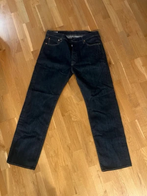 Aldrig använda Levis 501 - Snygga mörkblå jeans med rak passform och klassisk femficksdesign. Jeansen har kontrastsömmar och är tillverkade i ett robust denimtyg som ger en tidlös look. Perfekta för dig som gillar enkel och stilren stil. Jeansen är i storlek W36 L32 och aldrig använda, nypris ca 1000 kr.