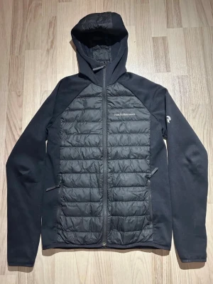 Svart dunjacka från Peak Performance - Snygg marinblå dunjacka från Peak Performance med huva och dragkedja framtill. Jackan har quiltade paneler på bröst, rygg och huva samt släta ärmar. Två dragkedjeförsedda fickor på sidorna och diskret logga på bröstet. Perfekt för kalla vinterdagar.