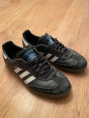 Adidas Samba - Ett par Adidas Samba sneakers i svart läder med vita detaljer. Skorna har svart mocka vid tån, svart snörning och mörkbrun gummisula. De är lite lätt skrapade vid hälen, annars inga defekter. 