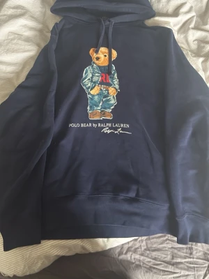 Marinblå hoodie Polo Bear Ralph Lauren - Säljer en marinblå hoodie från Ralph Lauren med den ikoniska Polo Bear-trycket på bröstet. Hoodien har dragsko i huvan och en klassisk känguruficka framtill. Materialet är mjukt och bekvämt, perfekt för en avslappnad stil.