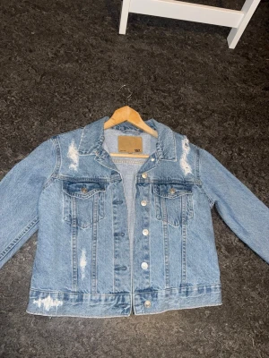 Ljusblå distressed jeansjacka 157 - Säljer en ljusblå jeansjacka från 157 med slitna detaljer och klassisk krage. Jackan har två bröstfickor med lock och knappar framtill. Perfekt oversized look och riktigt snygg vintage-vibe.