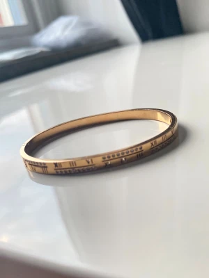 Guldfärgat armband med romerska siffror - Snyggt guldfärgat armband i metall med ingraverade romerska siffror runt hela bandet. Armbandet har även små gnistrande stenar infällda på ena sidan för extra bling. Perfekt accessoar för dig som gillar stilrena detaljer och en touch av lyx.