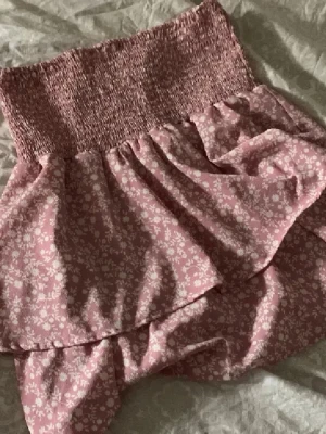 Rosa blommig volangkjol - Supersöt rosa kjol med blommigt mönster och volangdetaljer. Kjolen har en bred, smockad midja som ger en bekväm och flexibel passform. Går även att använda som topp