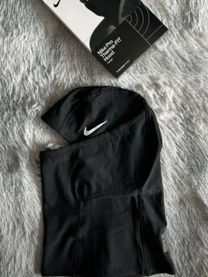 Nike Pro Therma-FIT svart balaclava - Nike Pro Therma-FIT balaclava i svart med vit Swoosh-logga på sidan. Tillverkad i ett tekniskt, stretchigt material som håller värmen och transporterar bort fukt. Sitter tight runt huvudet och halsen, perfekt för träning utomhus eller kalla dagar.