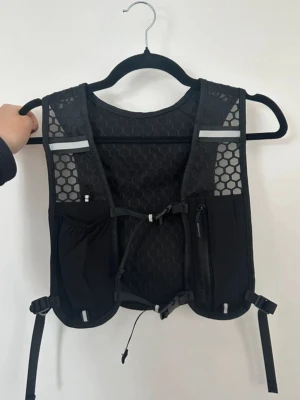 Svart löparväst med meshdetaljer - Säljer en svart löparväst med hexagonmönstrat meshmaterial för extra ventilation. Västen har flera fickor framtill och baktill, justerbara remmar och reflekterande detaljer. Perfekt för löpning eller trailrunning när du vill bära med dig småsaker. Onesize och unisexmodell.
