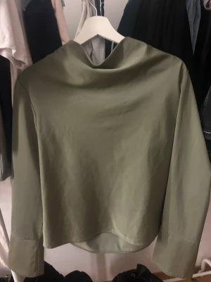 Olivgrön blus med draperad krage - Stilren olivgrön blus med lång ärm och unik draperad krage. Blusen har en lätt glansig yta och rak passform. Tillverkad i ett tunt, mjukt material som ger en elegant look. Perfekt för dig som gillar minimalistisk stil.