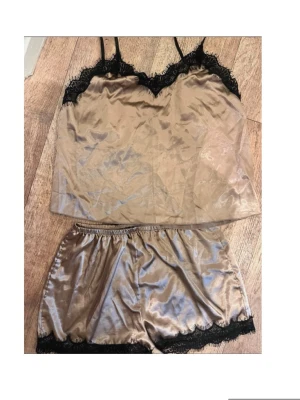 Champagnefärgat satinset med spets - Snyggt tvådelat set i champagnefärgad satin med svarta spetsdetaljer. Setet består av ett linne med tunna axelband och V-ringning samt matchande shorts med elastisk midja. Båda delarna har dekorativ svart spets längs kanterna för en extra lyxig känsla.
