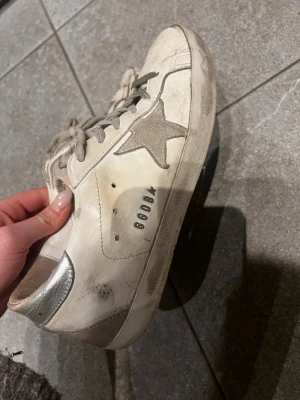 Golden Goose vita sneakers med stjärna - Golden Goose sneakers i vitt läder med grå stjärna på sidan och silverfärgad häl. Skorna har grå snörning och en klassisk låg siluett. Ikonisk GGDB-detalj på sidan och slitna detaljer för en trendig look.