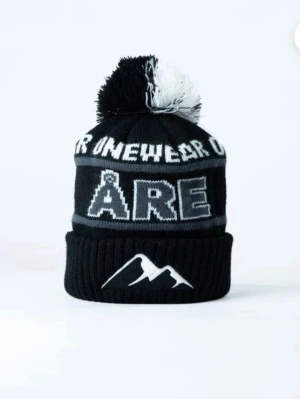 Svart Åre-mössa  - Säljer en svart och vit ribbstickad mössa från Onewear med stor tofs på toppen. Mössan har texten 'ÅRE' framtill och ett stiliserat bergmotiv på uppviket. Perfekt för dig som gillar streetstyle och vill ha en varm accessoar med cool design.