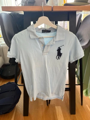 Ljusblå pikétopp från Polo Ralph Lauren - Ljusblå pikétopp från Polo Ralph Lauren med klassisk krage, korta ärmar och två knappar framtill. Stor mörkblå broderad logga på bröstet ger en sportig vibe. Tillverkad i mjuk bomull för skön känsla.