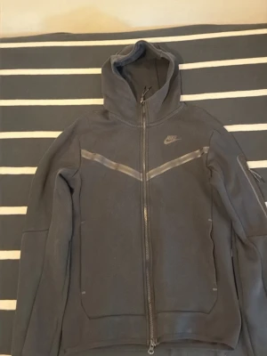Svart Nike hoodie med dragkedja - Snygg svart hoodie från Nike med hel dragkedja framtill och huva. Hoodien har diskreta reflexdetaljer över bröstet och en liten Nike-logga på vänster sida. Tillverkad i mjukt material som känns bekvämt mot huden. Perfekt för en sportig och avslappnad stil.