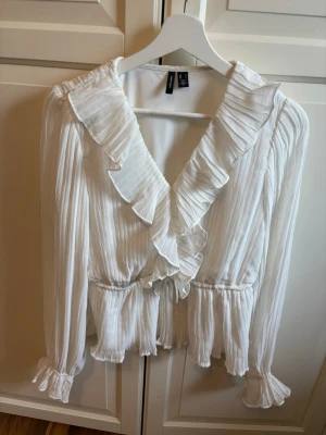Vit plisserad blus med volang H&M - Snygg vit plisserad blus från H&M med volangkant längs ringningen och knytband i midjan. Blusen har långa ärmar med resår och volang vid ärmslut samt en lätt glansig finish. Perfekt för en trendig och feminin look.