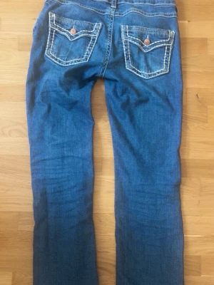 Blå bootcut jeans med kontrastsömmar - Säljer ett par klassiska blå bootcut jeans med vita kontrastsömmar och två bakfickor med dekorativa knappar. Jeansen har normal midja och stängs med knapp och dragkedja. Perfekta för en avslappnad och trendig look.
