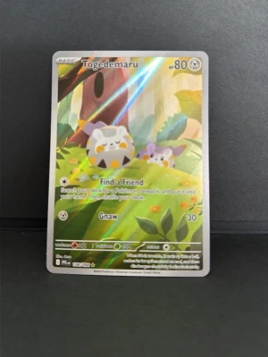 Togedemaru holografiskt Pokémonkort - Holografiskt Pokémonkort med Togedemaru och färgglad naturbakgrund. Kortet har glansig yta och klassisk blå baksida med Pokéball. Perfekt för samlare eller dig som gillar Pokémonkort.