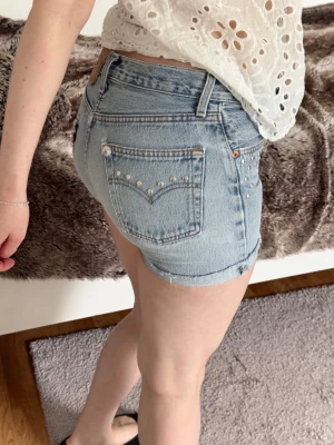 Levi's ljusblå denim shorts med nitar - Säljer ett par ljusblå denim shorts från Levi's med coola nitar på bakfickorna. Shortsens klassiska femficksdesign och råa benslut ger en avslappnad vibe. Perfekta för sommarens alla äventyr!