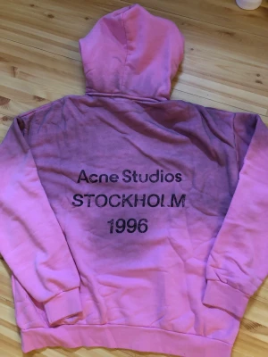 Rosa hoodie Acne Studios Stockholm - Säljer en rosa hoodie från Acne Studios med tryck på ryggen där det står 'Acne Studios STOCKHOLM 1996'. Hoodien har en klassisk huva och ribbade muddar vid ärmar och nederkant. Perfekt för dig som gillar streetwear och vill ha något unikt från ett svenskt märke.