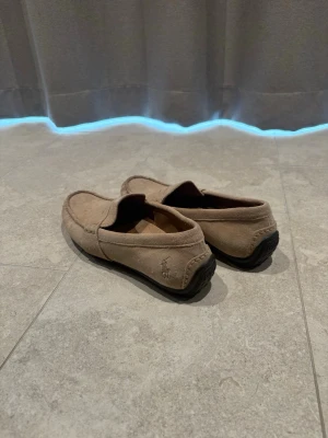 Beige loafers från Polo Ralph Lauren - Snygga beige loafers i mocka från Polo Ralph Lauren med klassisk design och svart sula. Perfekta för en avslappnad men stilren look. Ikoniska Polo-loggan syns på sidan och insidan av skon.