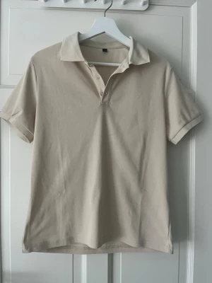 Supersnygg t shirt - Klassisk beige pikétröja med korta ärmar och krage. Tröjan har knappar vid halsen och är tillverkad i ett mjukt bomullsmaterial. Enkel och stilren design som passar till många olika looks.
