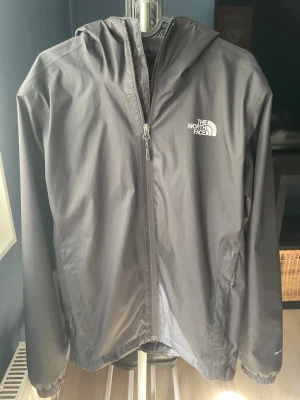 Svart regnjacka från The North Face - Snygg svart regnjacka från The North Face med huva och dragkedja framtill. Jackan har två sidofickor med dragkedja och justerbar nederkant. Tillverkad i ett vattenavvisande material som håller dig torr under regniga dagar. Perfekt för dig som vill ha en stilren och funktionell jacka.