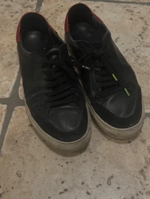 Svarta Arigatos låga sneakers med röd detalj - Säljer ett par svarta sneakers med lågt skaft och röd detalj vid hälen. Skorna har svarta snören och vit sula. Perfekta för dig som gillar en klassisk och stilren look med en liten färgklick.