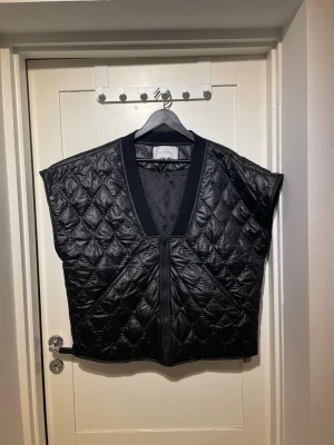 Svart Quiltad Copenhagen Muse jacka med avtagbara ärmar - Svart quiltad jacka med dragkedja framtill samt avtagbara ärmar så den kan bäras som väst. Jackan har V-ringad hals och ett glansigt material som ger en modern look. Perfekt för lager på lager och enkel att styla till olika outfits. Givetvis kan priset diskuteras.