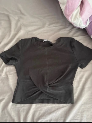 Svart croppad topp med twistdetalj - Snygg svart croppad topp med twistdetalj framtill. T-shirten har rund halsringning och korta ärmar. Perfekt till jeans eller kjol för en trendig look.