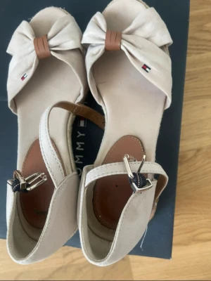 Beige kilklackade sandaler Tommy Hilfiger - Passar 36/37 Snygga beige sandaler från Tommy Hilfiger med kilklack i flätad jute och öppen tå. Framtill har de en stor rosett och små logodetaljer. Rem runt ankeln med spänne i metall. Perfekta för sommarens alla outfits.