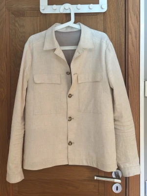 Beige linneskjorta overshirt - Stilren beige overshirt i linne med klassisk krage och fyra knappar framtill. Två stora bröstfickor med lock ger en avslappnad look. Perfekt lager på lager-plagg för dig som gillar minimalistisk stil. (Dock är en knapp trasig men funkar fortfarande o knäppa)