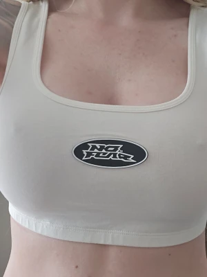 Beige linnetopp från No Fear x H&M - Säljer en beige linnetopp från No Fear med breda axelband och ett stort logomärke framtill. Toppen har en croppad passform och är tillverkad i mjuk bomull, perfekt för en avslappnad och sportig stil.