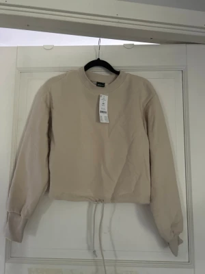 Beige cropped sweatshirt från H&M - Säljer en beige cropped sweatshirt från Gina tricot med långa ärmar och dragsko i nederkanten. Tröjan har en enkel och clean design utan huva, rund halsringning och ribbade muddar. Tröjan är lite kortare i modellen. Snörning i nedre delan av tröjan. Säljer den pågrund av att den har blivit förliten🥰