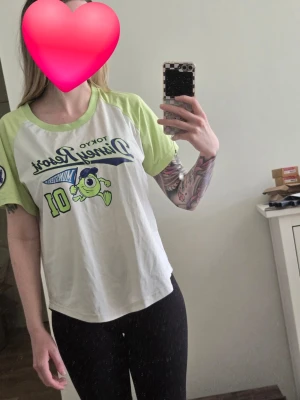 Tokyo Disney Resort Monsters U T-shirt - Säljer en vit och ljusgrön T-shirt från Tokyo Disney Resort med Monsters University-tryck och Mike Wazowski på framsidan. T-shirten har kontrastfärgade ärmar och krage samt coolt tryck i blått och grönt. Perfekt för dig som gillar Disney och streetstyle. Köpt i Tokyo 2017