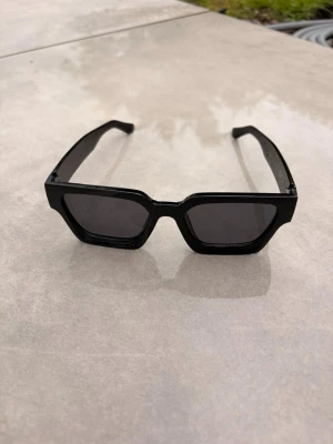 Svarta kantiga solglasögon - Snygga svarta solglasögon med breda, kantiga bågar och mörka glas. De har en modern och edgy look som passar perfekt till en trendig stil. Ramen är i plast och designen är riktigt statement.🕶️☀️⛱️