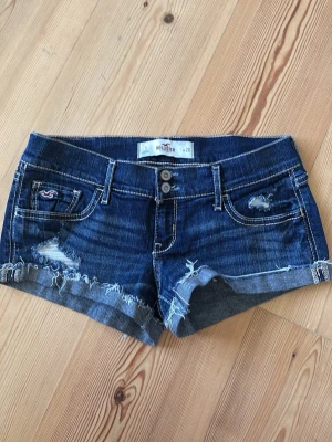Mörkblå jeansshorts från Hollister - Snygga mörkblå jeansshorts från Hollister med slitna detaljer och råa kanter. Klassisk femficksmodell med dubbla knappar framtill och dekorativa sömmar på bakfickorna. Perfekta för en avslappnad och trendig look. storlek w26, slutsåld 🩷skulle säga storlek 34/XS-S eller samma som w26 i true religion och weekday 💕