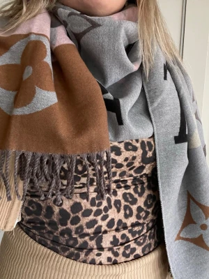 Vuitton mönstrad halsduk - Snygg halsduk från Vuitton i mjukt material med stora monogram och blommönster. Färgerna är grå, brun och beige med inslag av ljusrosa. Halsduken har fransar i ändarna och är perfekt för att lyfta din outfit cashmire 