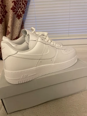 Nike Air Force 1 '07 Vit - Klassiska Nike Air Force 1 '07 sneakers i helvitt läder med vit sula och vita skosnören. Ikonisk swoosh på sidan och AIR-detalj på hälen. Stilren och tidlös modell som passar till det mesta. Perfekt för dig som gillar clean och enkel stil.