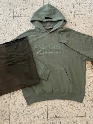 Essentials Fear Of God Hoodie- grön/turkos - Snygg grön hoodie från Fear of God Essentials med stor logga på bröstet. Riktigt unik färg som kommer få dig att sticka ut. Storlek S. Original tags och påse medföljer.