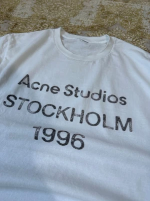 Vit Acne Studios t-shirt med tryck - Vit t-shirt från Acne Studios med svart tryck på bröstet där det står 'Acne Studios STOCKHOLM 1996'. Klassisk rund halsringning och korta ärmar. Perfekt för en clean och stilren look.
