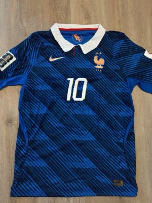 Blå Frankrike Nike fotbollströja Mbappe - Säljer en blå fotbollströja från Nike med Frankrikes landslagsemblem och nummer 10 på bröstet. Tröjan har vita detaljer, två stjärnor ovanför emblemet och en vit krage. Tillverkad i lätt och ventilerande material, perfekt för match eller träning.