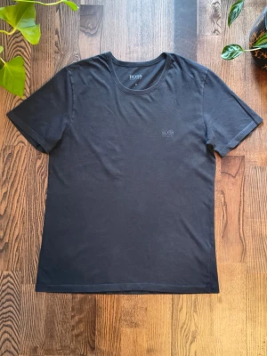 Svart t-shirt från Hugo Boss - Klassisk svart t-shirt från Hugo Boss med diskret broderad logga på bröstet. T-shirten har rund halsringning och korta ärmar. Tillverkad i mjuk bomull för en skön känsla och enkel stil.