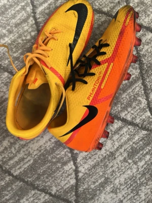 Nike Phantom GT fotbollsskor gul/orange - Säljer ett par Nike Phantom GT fotbollsskor i en riktigt snygg gul och orange färg med svarta detaljer och logga. Skorna har dobbar för bra grepp på planen och är tillverkade i syntetmaterial som ger bra bollkontroll. Perfekta för dig som vill sticka ut på fotbollsplanen!