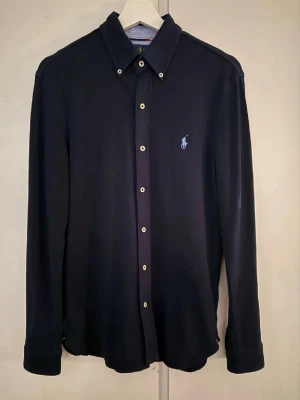 Mörkblå skjorta från Ralph Lauren - Klassisk mörkblå skjorta från Ralph Lauren med knappslå och ikoniska broderade loggan på bröstet. Skjortan har långa ärmar, vita knappar och en tidlös design som passar till det mesta. Perfekt för dig som gillar stilrena plagg säljer den då den ej kommer till användning kan även gå ner i pris vid snabb affär.