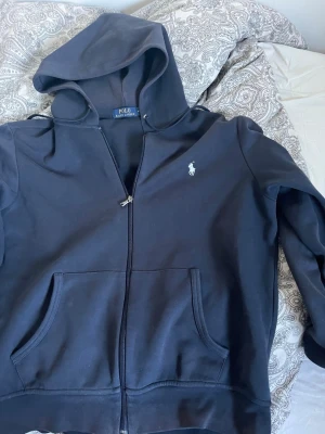 Svart hoodie från Polo Ralph Lauren - Snygg svart hoodie från Polo Ralph Lauren med dragkedja framtill och klassisk vit logga på bröstet. Hoodien har huva, två stora fickor och är gjord i mjukt bomullsmaterial. Perfekt för en chill och stilren look.