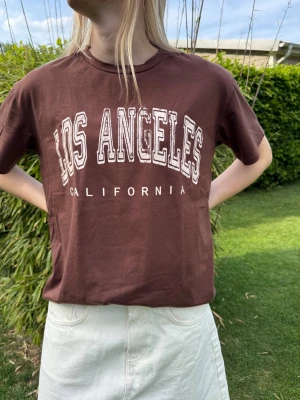 Brun Los Angeles t-shirt - Snygg brun t-shirt med trycket 'Los Angeles California' i vitt framtill. Klassisk passform med rund hals och korta ärmar. Perfekt till jeans eller kjol för en avslappnad look.