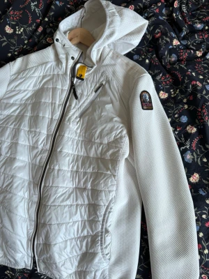 Vit hybridjacka från Parajumpers - Snygg vit hybridjacka från Parajumpers med quiltad front, huva och hel dragkedja. Jackan har en bröstficka med dragkedja och klassisk PJS-logga på ärmen. Perfekt för lager-på-lager och kyliga dagar.