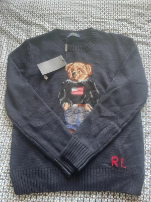 Mörkblå stickad tröja Polo Bear - Mörkblå stickad tröja från Ralph Lauren med den ikoniska Polo Bear på bröstet, klädd i jeans och tröja med amerikanska flaggan. RL-broderi i rött vid nederkanten. Rund halsringning och ribbade muddar. Perfekt för dig som gillar klassisk preppy stil.