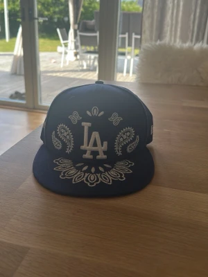 Blå LA keps med bandanamönster - Säljer en blå New Era keps med LA-logga framtill och vitt bandanamönster både fram och bak. Kepsen har rak skärm och klassisk snapback-modell. Perfekt för dig som gillar streetwear och vill sticka ut med snygga detaljer.